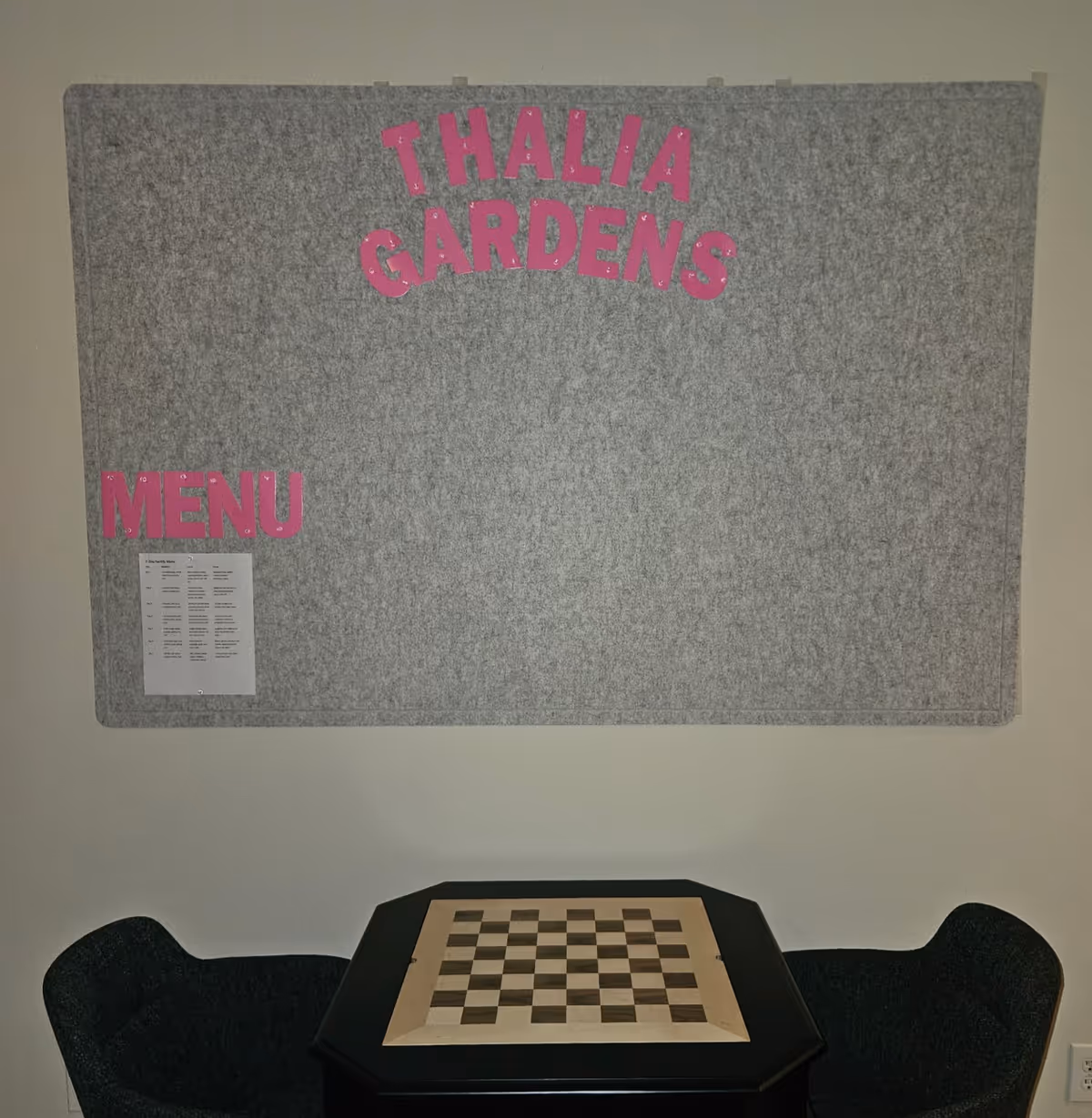 Grey bulletin board with pink 'Thalia Gardens' lettering above a chessboard table.