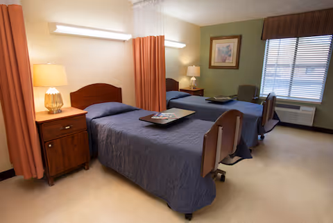 Shared senior living bedroom with two beds, curtains, lamps, and a window desk area.