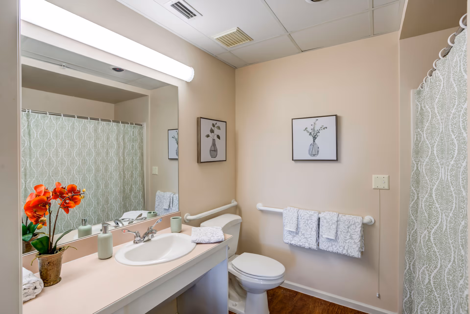 A clean bathroom with a white sink and countertop, a large mirror above the sink, a toilet with grab bars on the walls, a towel rack holding three patterned towels, a framed picture of a vase with greenery on the wall, a shower with a patterned curtain, and a small potted plant with orange flowers on the countertop.