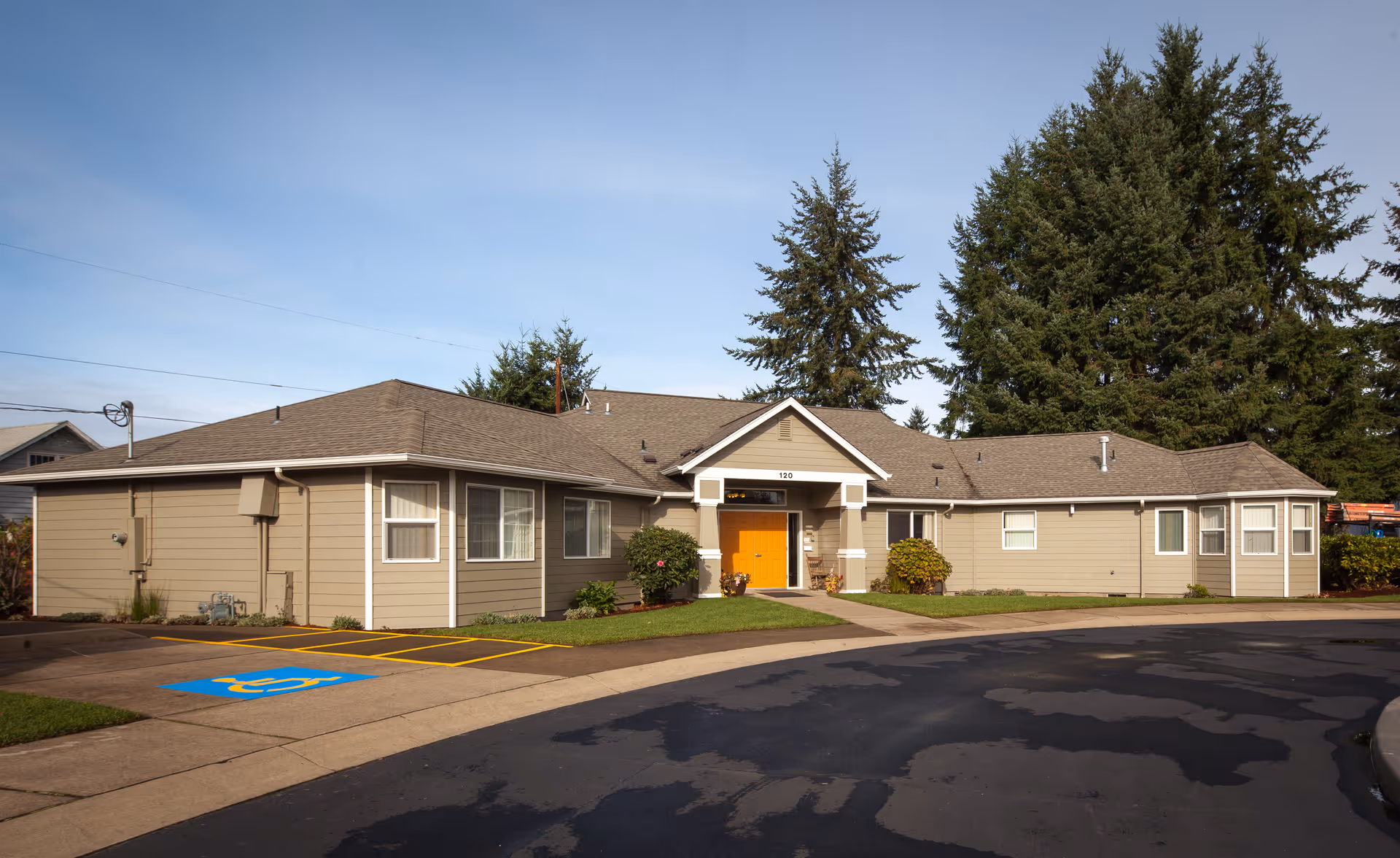 Exterior view of a single-story building with beige siding and a bright orange front door, surrounded by green grass and tall evergreen trees under a clear blue sky. There is a handicapped parking space in front of the building.