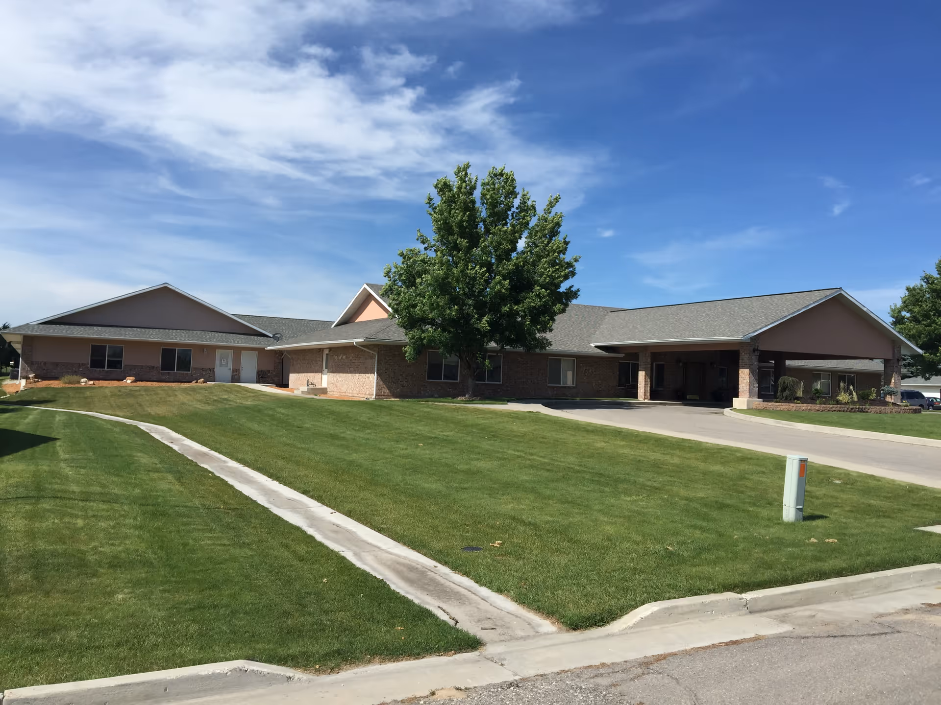 Exterior view of a single-story senior living facility building with a large green lawn and a tree in front under a partly cloudy blue sky.