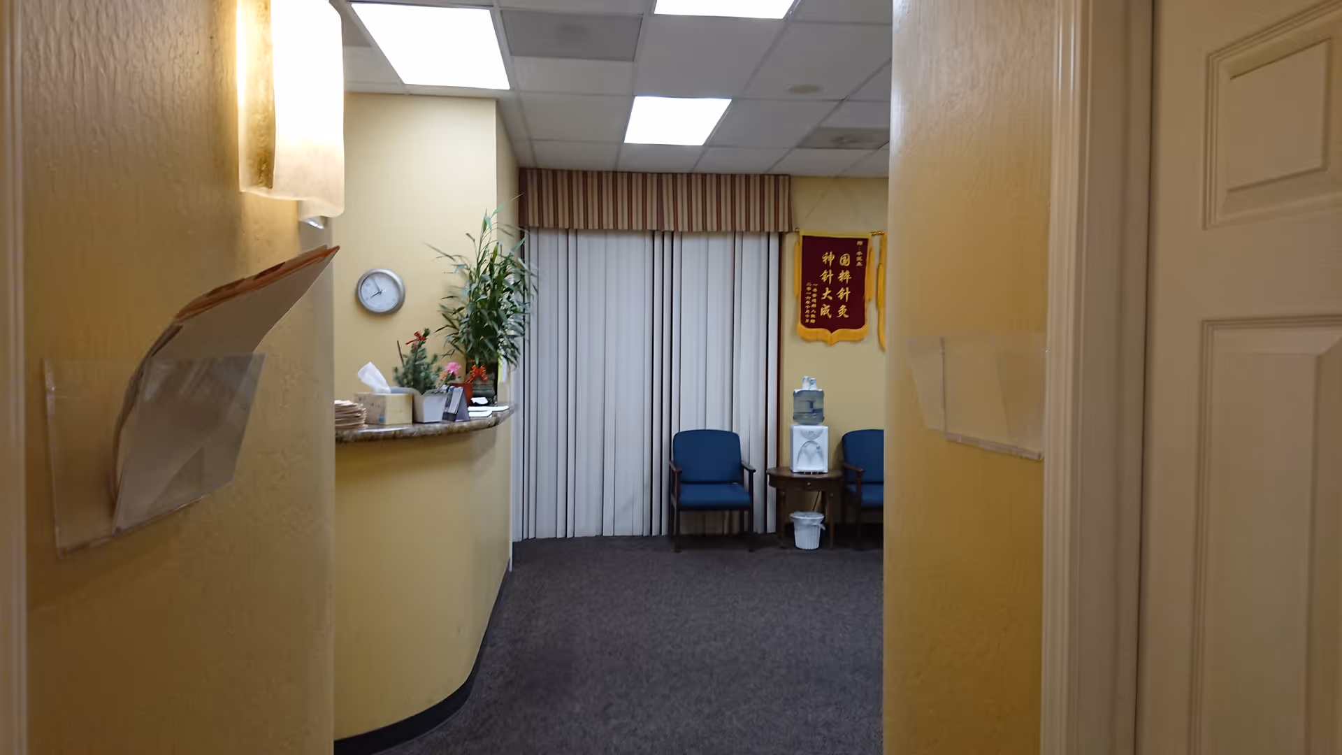 Reception area with curved counter, clock, plants, and blue chairs in a hallway.