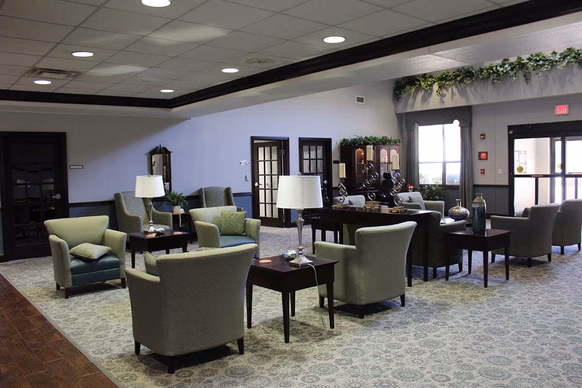 Spacious communal seating area with upholstered armchairs, side tables, lamps, and decorative plants in a senior living facility lobby.