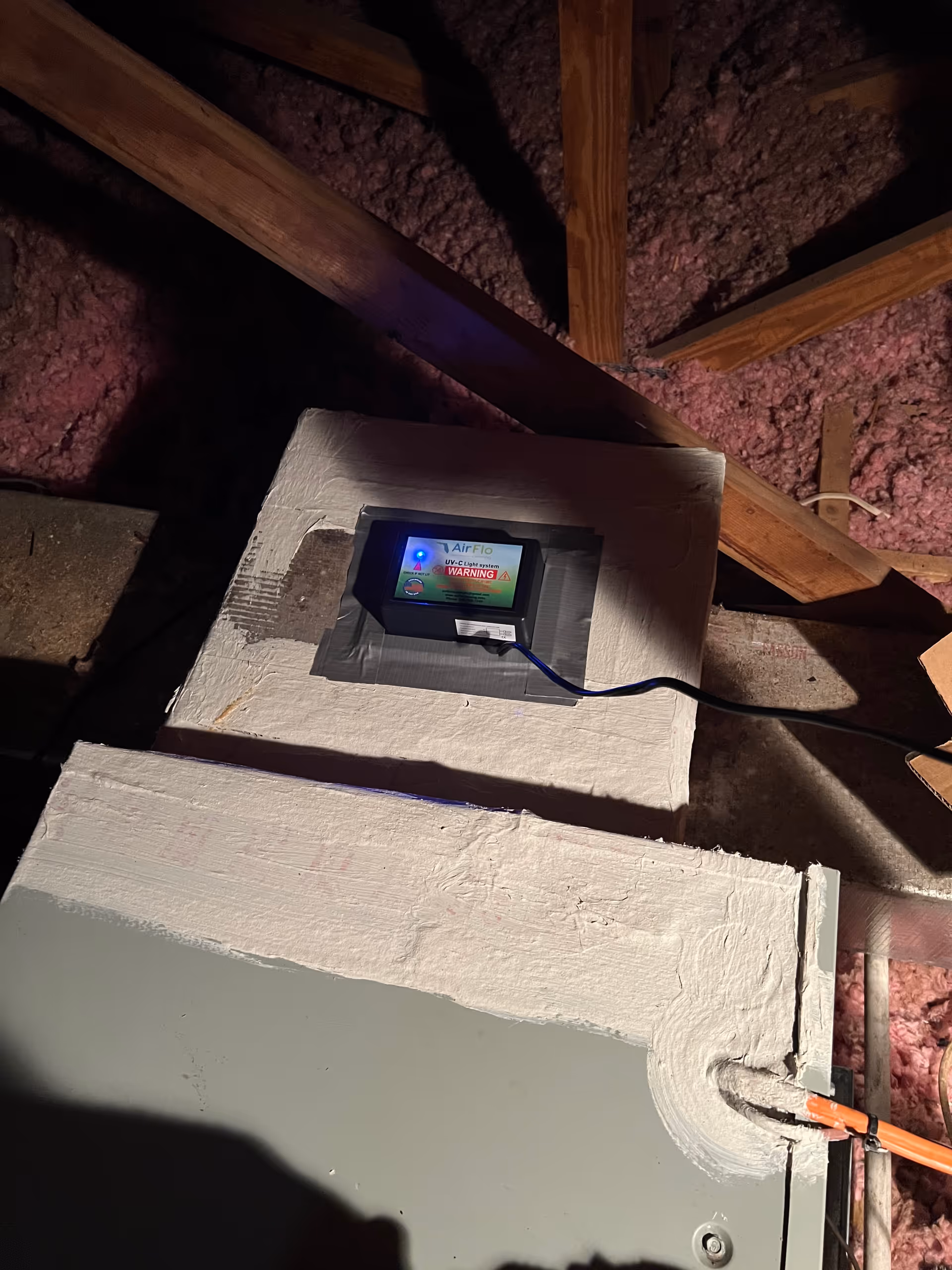 Attic space with pink insulation, wooden beams, and a mounted device displaying a screen.