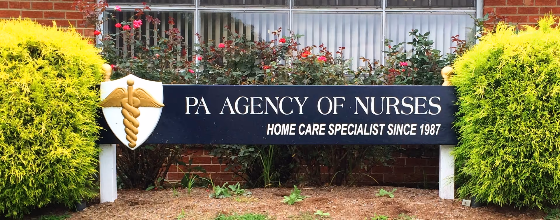 Exterior view of a PA Agency of Nurses sign with greenery and a brick building.
