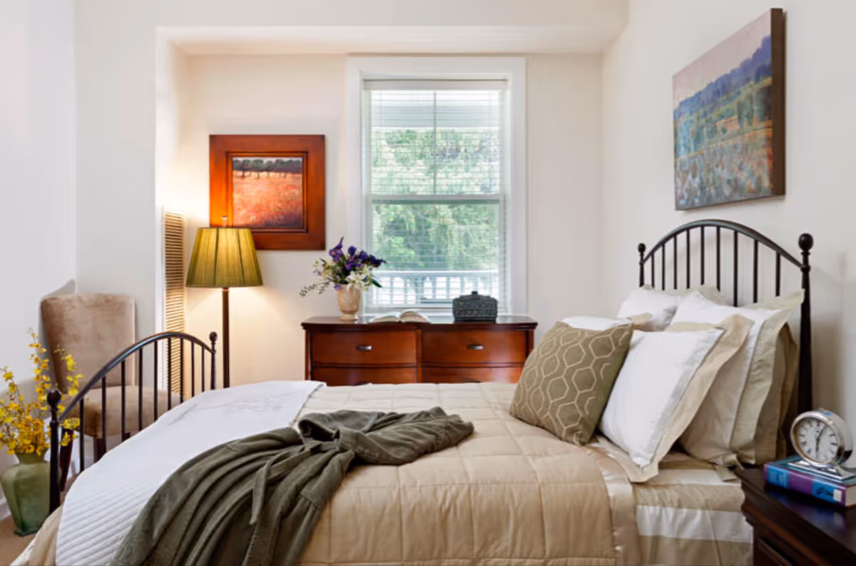 Sunny bedroom with a metal-framed bed dressed in beige bedding, a dresser beneath a window, a lamp, chair and wall art.