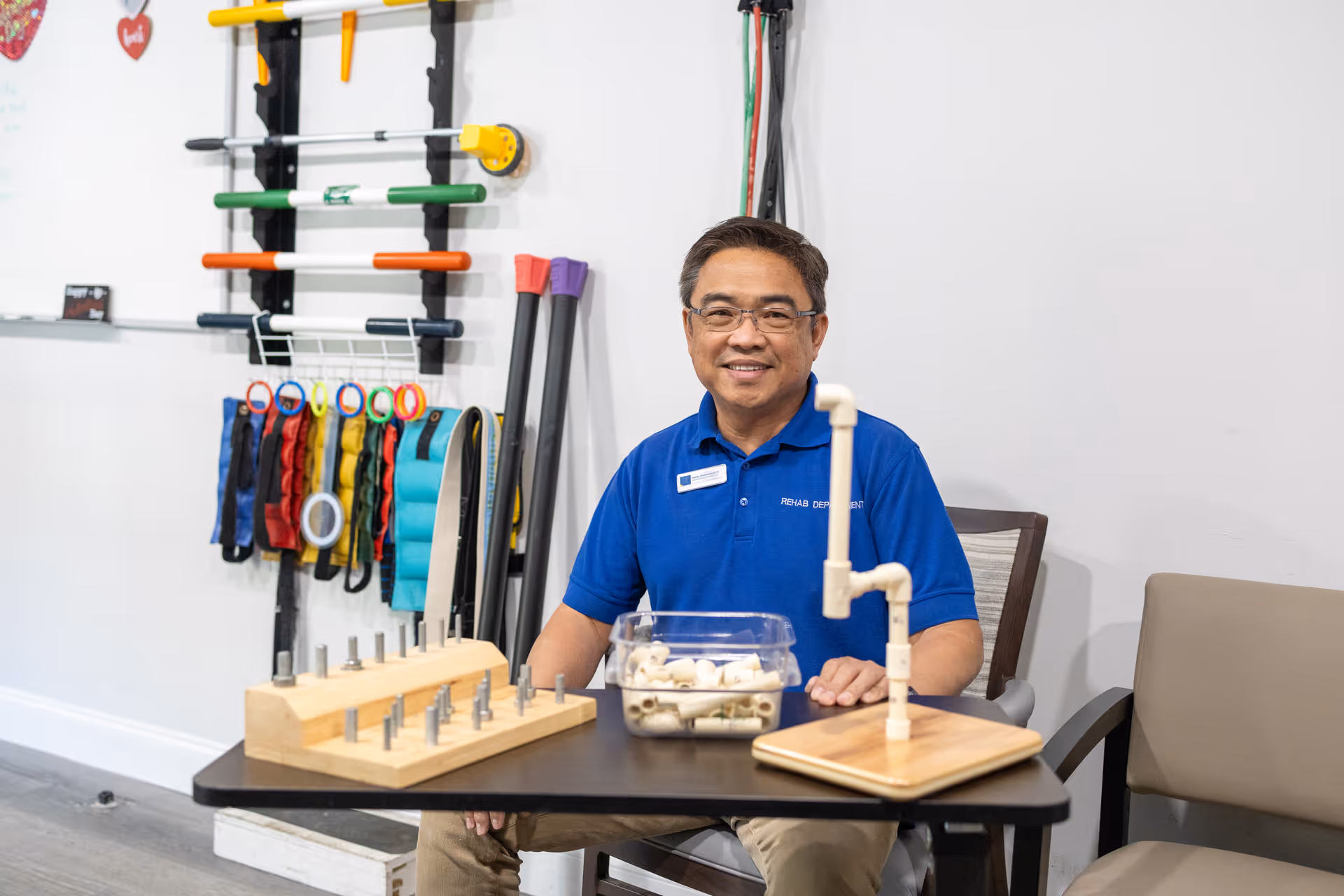 A man wearing glasses and a blue polo shirt with a Rehab Department badge sits at a table with various therapy tools and equipment in a room with exercise bands and rods hanging on the wall behind him.