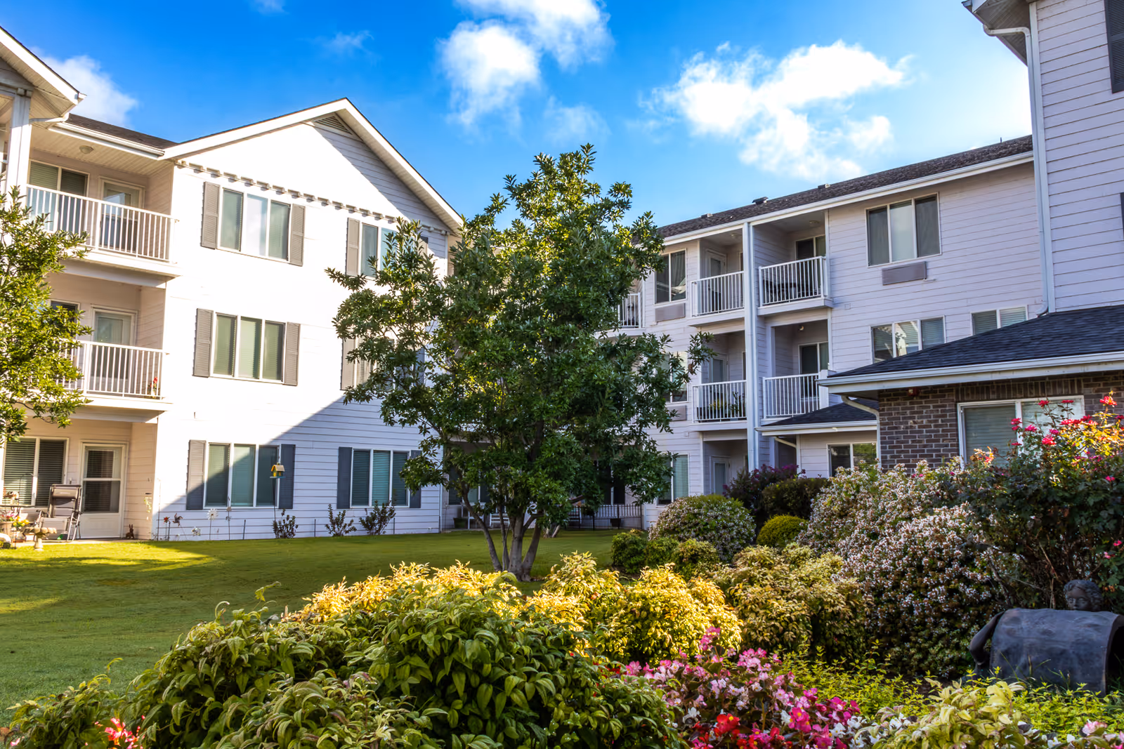 Exterior view of a senior living facility with white multi-story buildings surrounding a landscaped garden area featuring green bushes, flowering plants, and a tree under a blue sky with some clouds.