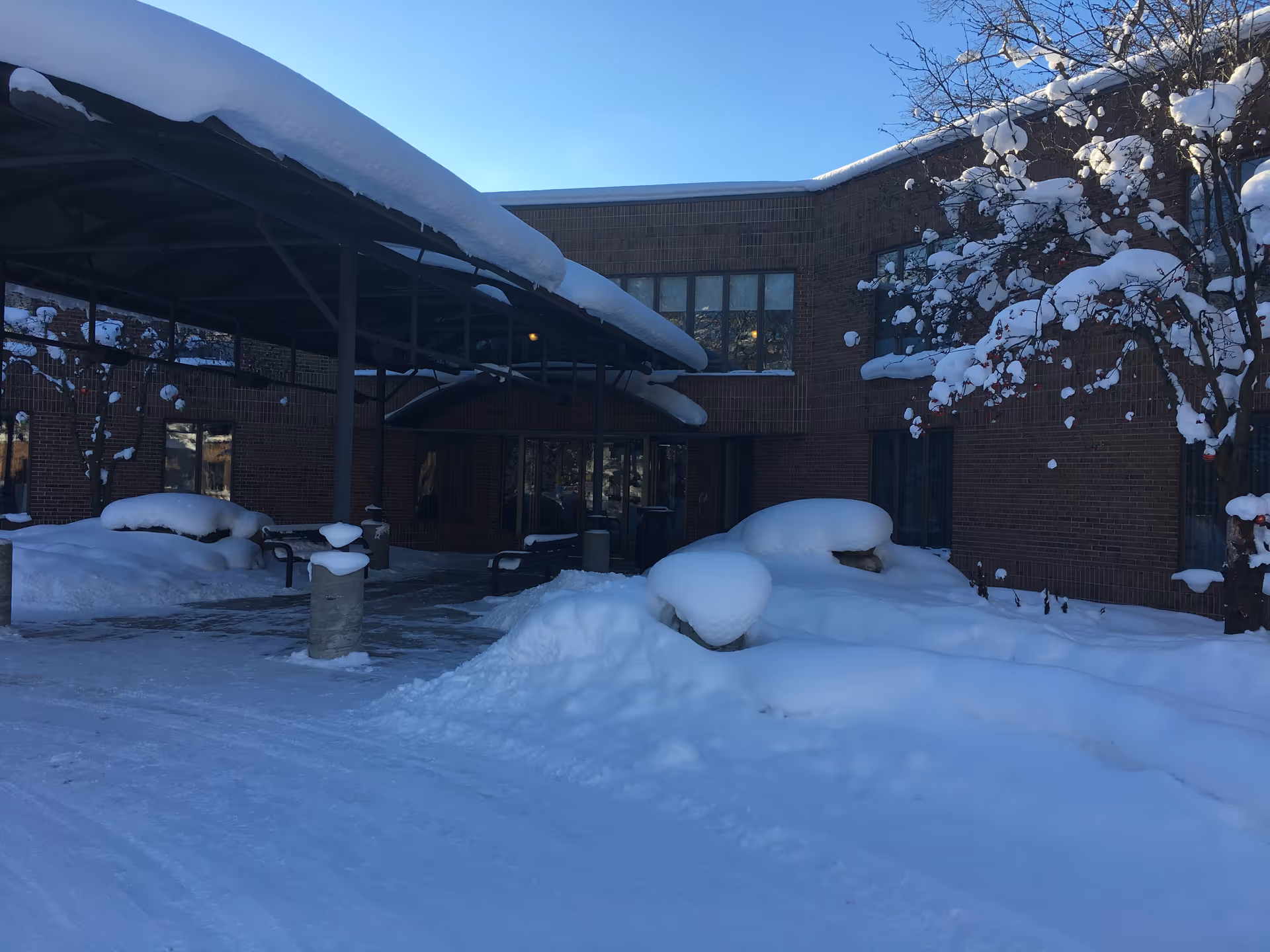The snow-covered exterior entrance of Robbinswood Northcrest shows a brick facade and snow-laden tree.