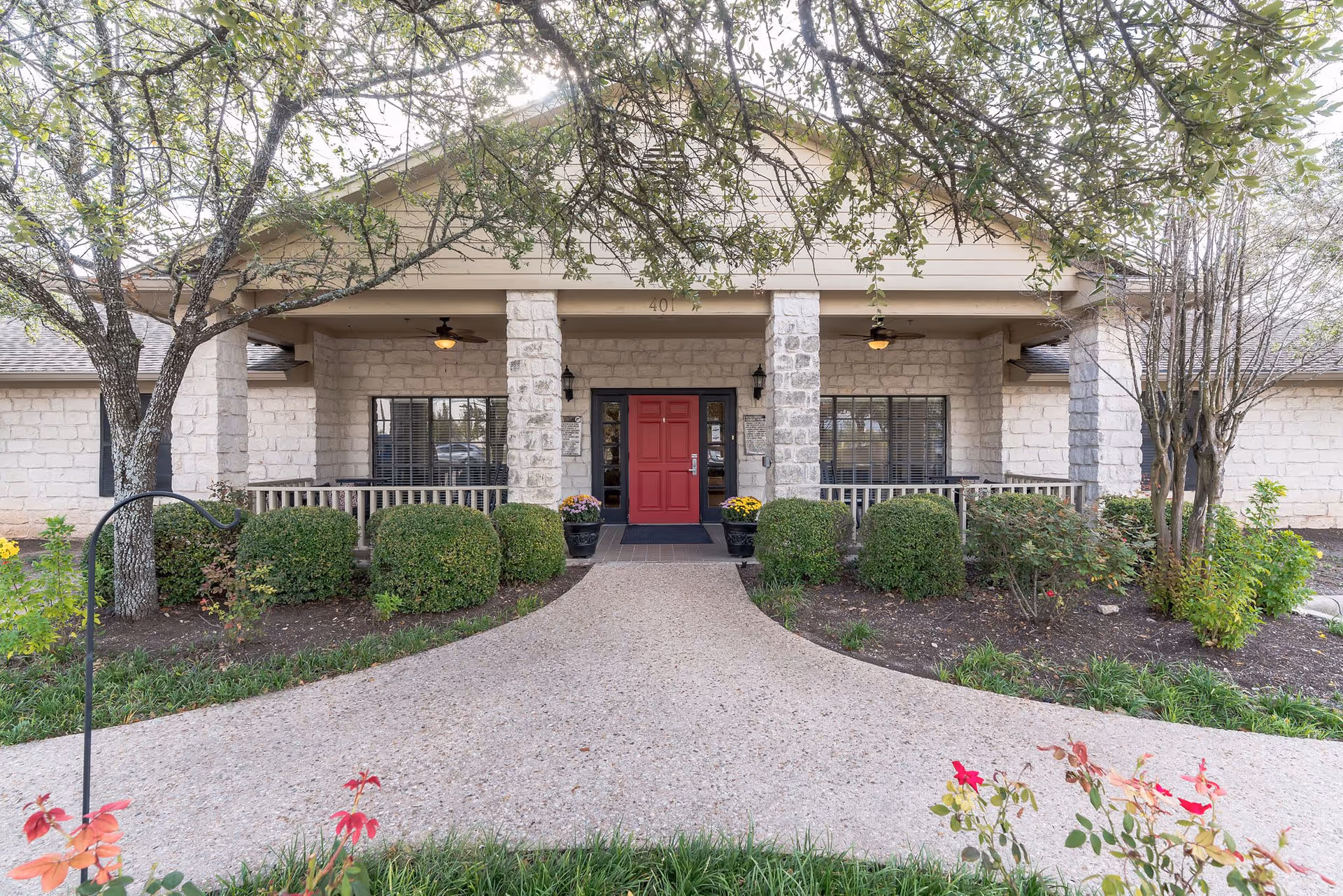 Front entrance of a stone-clad assisted living building with a covered porch, red door, columns, shrubs, and a curved walkway.