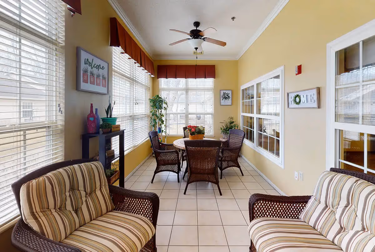 Sunlit sunroom with wicker armchairs and a round table surrounded by large windows and potted plants.