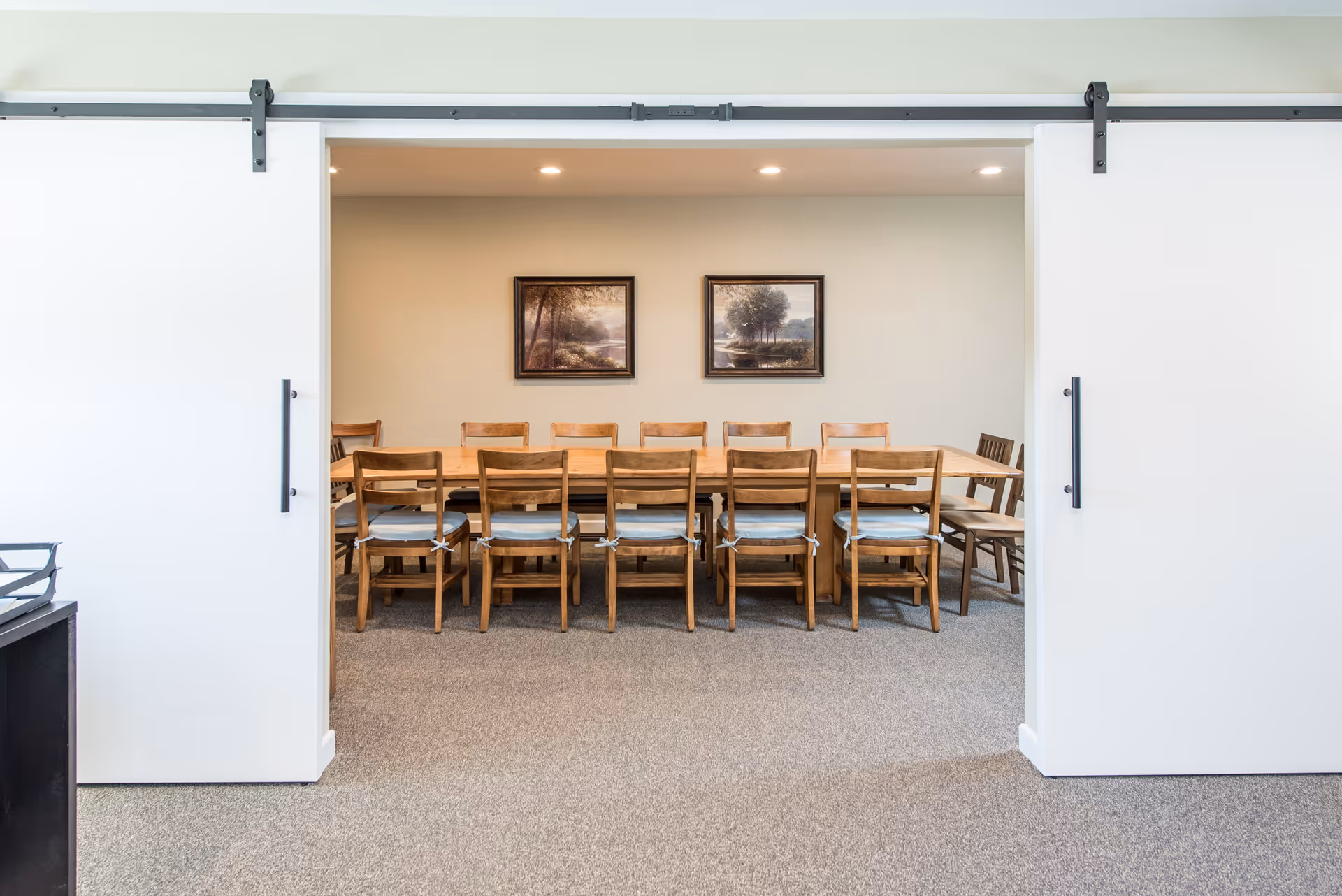 Sliding white barn doors reveal a dining room with a long wooden table, multiple chairs, and two framed landscape paintings on the back wall.