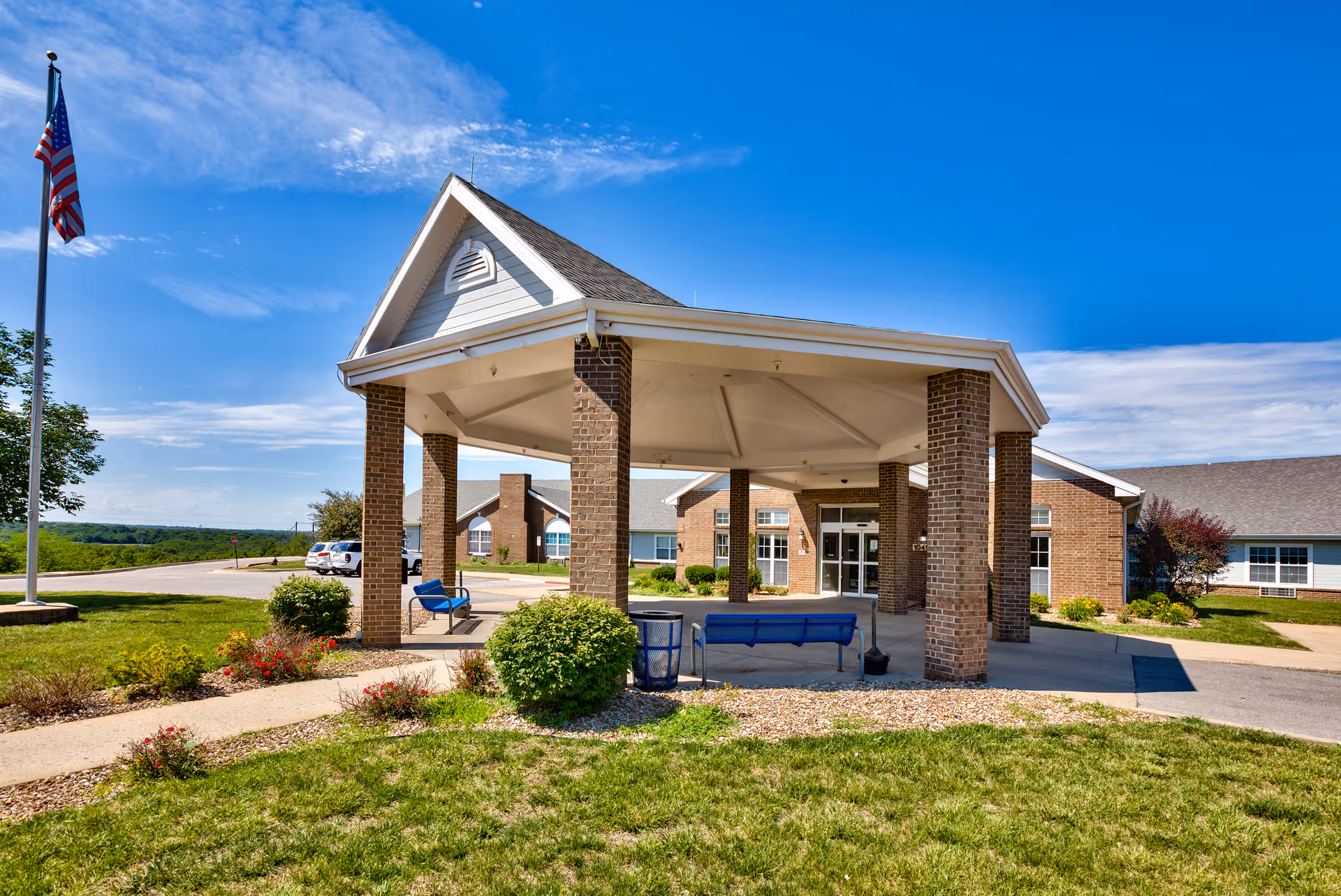 Front entrance with a covered porte-cochere of a brick senior living building, blue benches, landscaped grounds and an American flag.