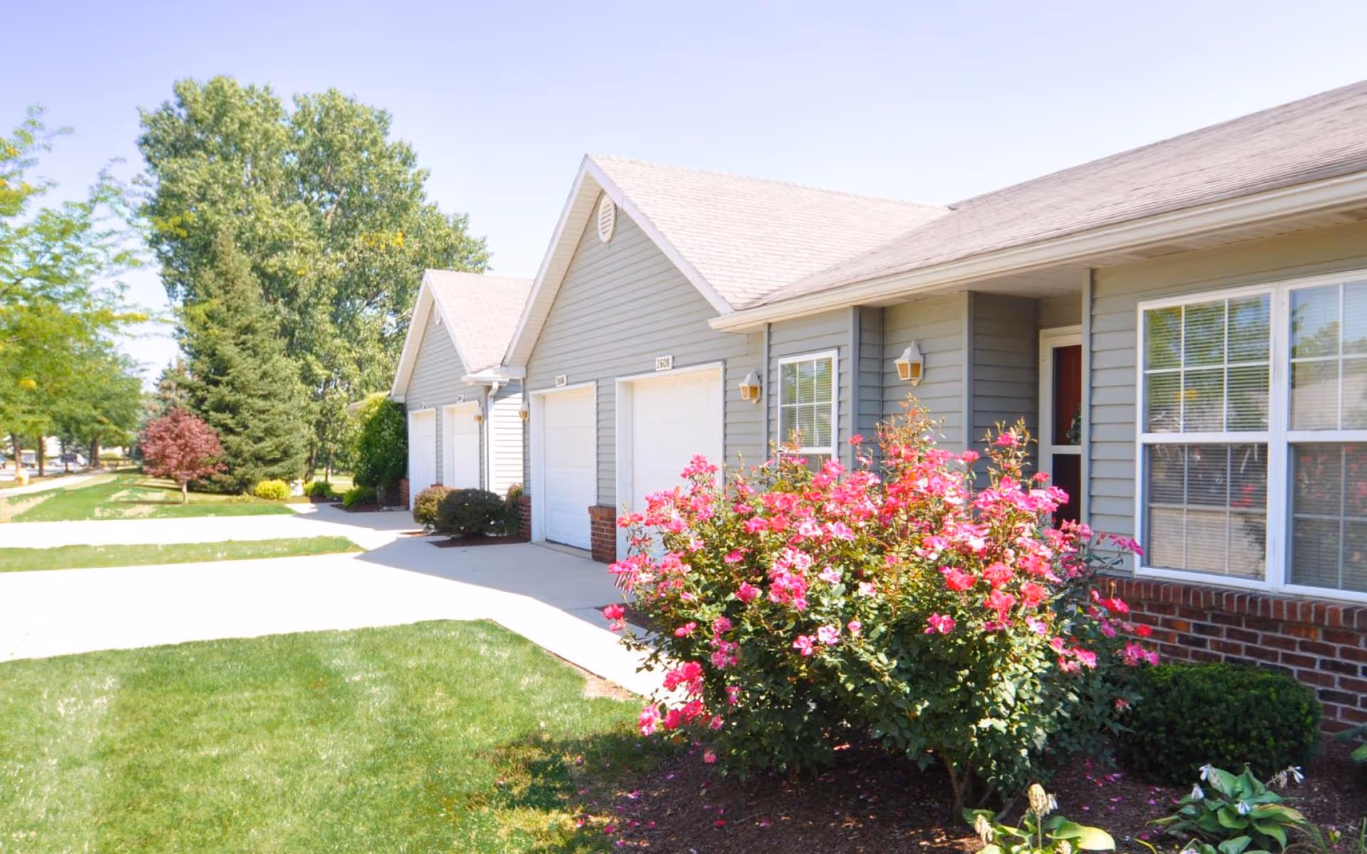 Exterior view of a residential building with gray siding and white garage doors. There is a well-maintained lawn and a vibrant pink flowering bush in front of the building. Trees and a sidewalk are visible in the background under a clear blue sky.