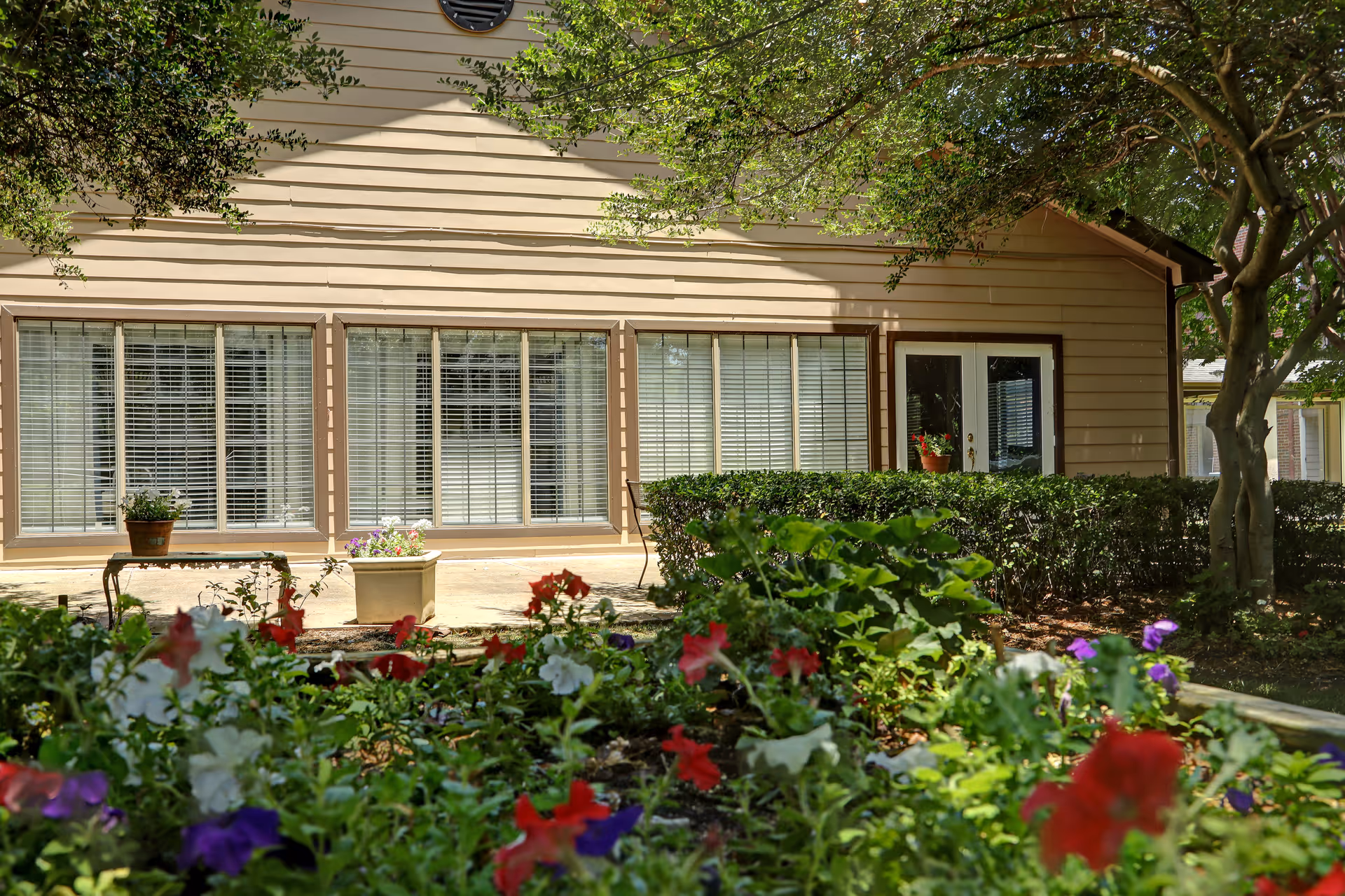Exterior view of a beige building with large windows and a glass door, surrounded by green bushes and colorful flowers in the foreground, with trees providing shade.