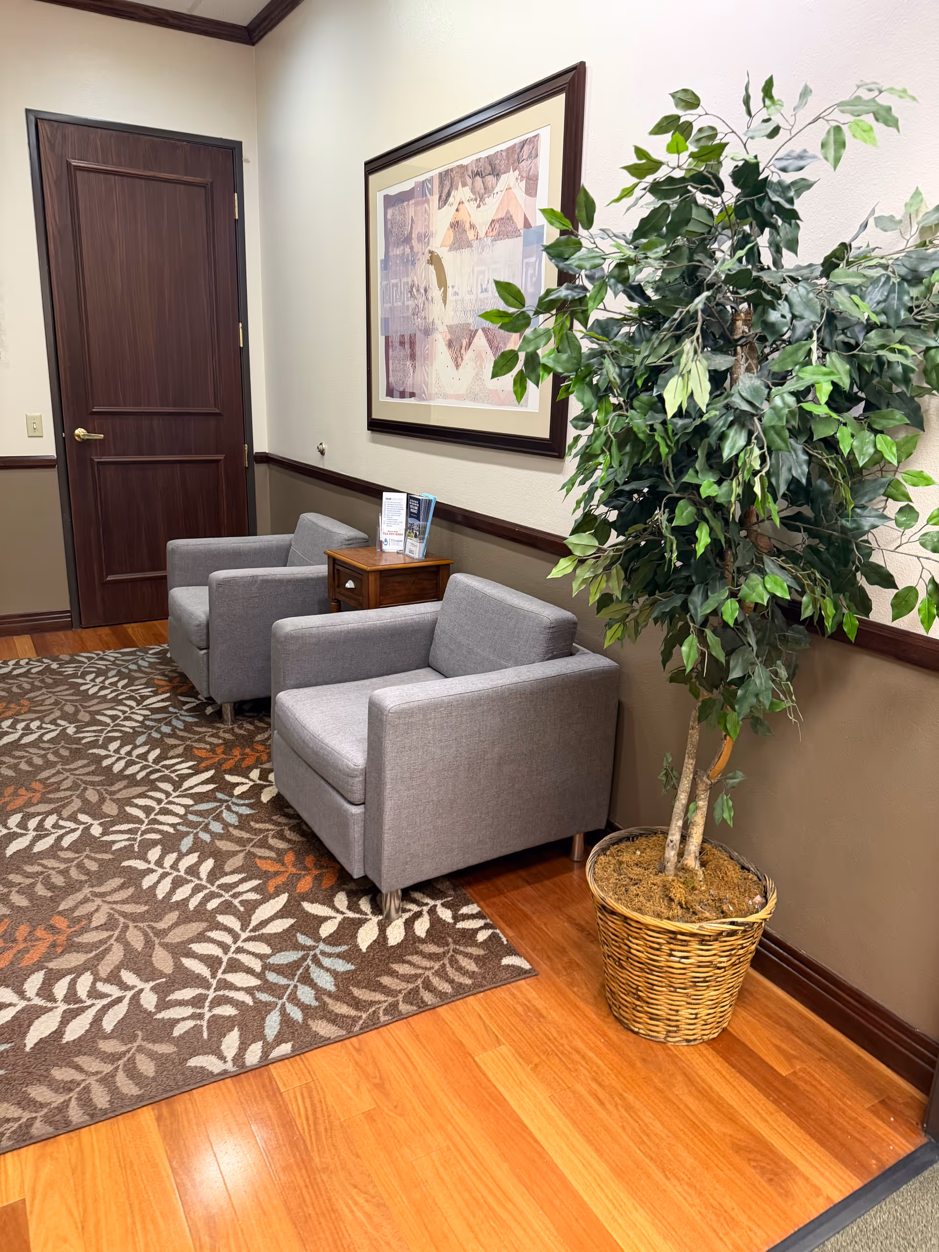 Waiting area with two gray armchairs, a side table, framed artwork, rug, and a potted plant.