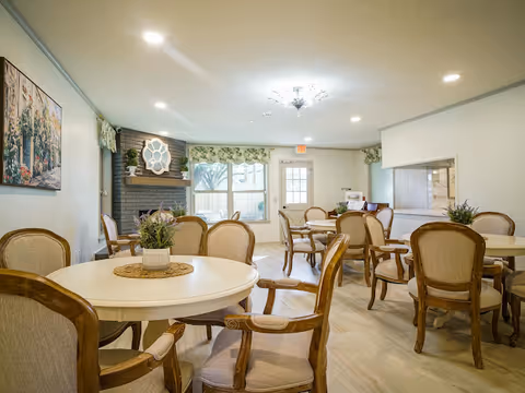 Bright, cozy dining room with round tables, beige chairs, a fireplace, and potted plants.