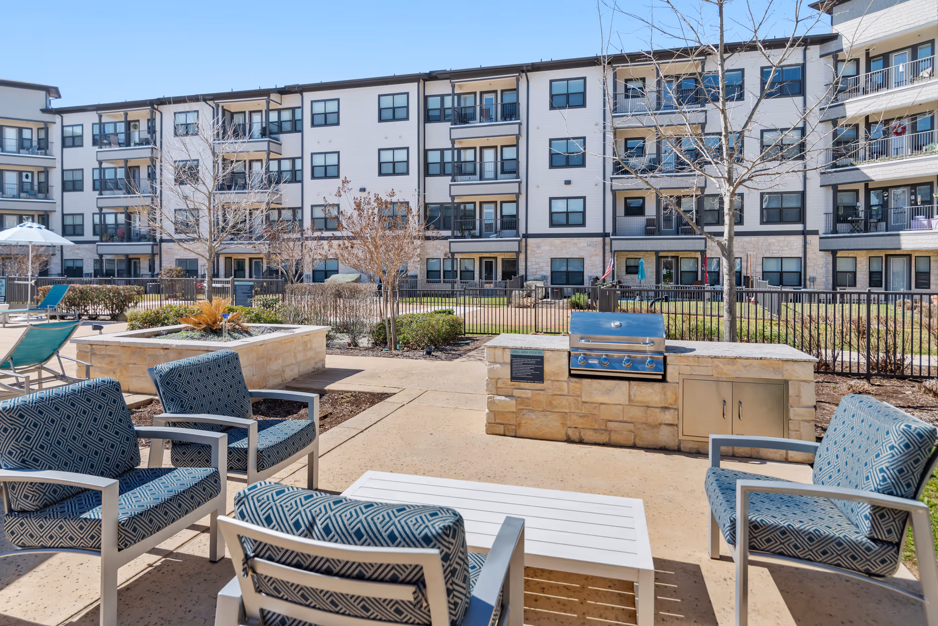 Outdoor patio with blue patterned chairs and a grill, facing a multi-story senior living building.