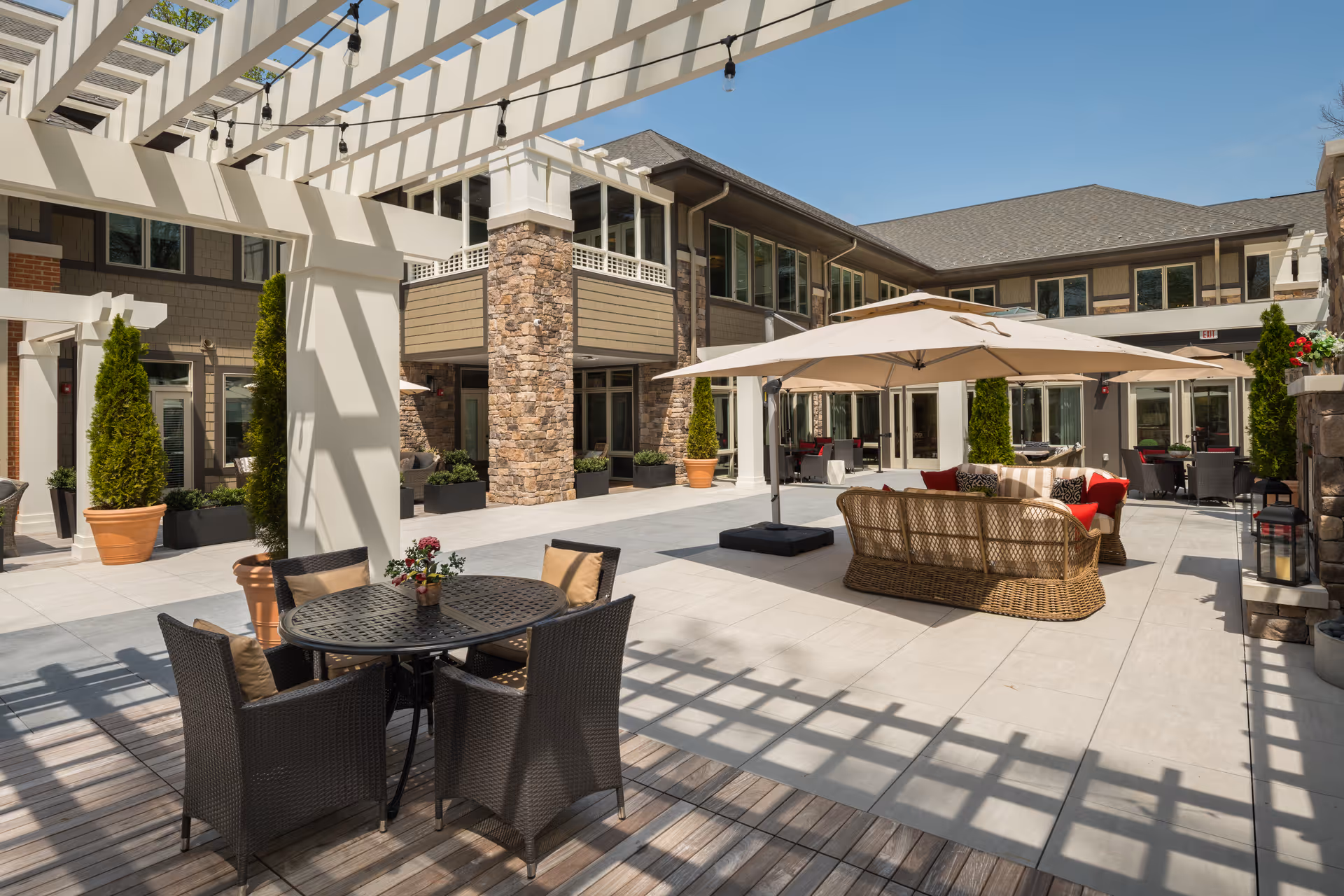 Sunny outdoor courtyard at The Kensington Reston featuring wicker sofas, umbrella-shaded seating, a dining table, and a pergola beside a two-story building.