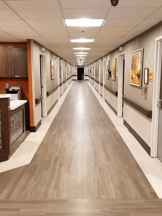 A long, well-lit interior hallway of a care facility with patient room doors and framed artwork along both walls.