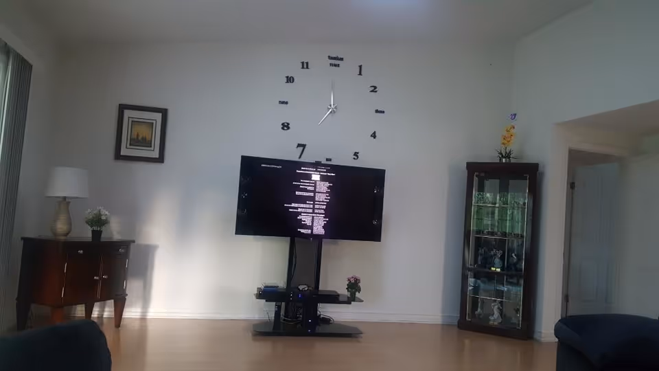 A living room with a large wall clock above a flat-screen TV mounted on a stand. To the left is a wooden side table with a lamp and a small plant, and to the right is a glass display cabinet with decorative items. The room has light-colored walls and wooden flooring.