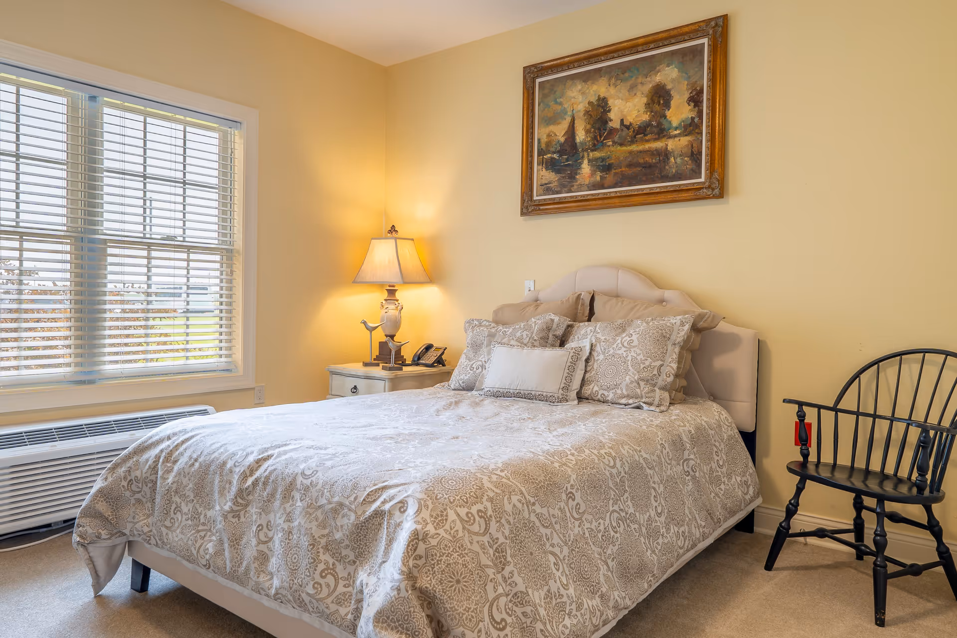 A cozy bedroom with a large bed covered in a patterned beige and white comforter and multiple pillows. There is a nightstand with a lamp and a phone next to the bed. A framed painting hangs on the yellow wall above the bed. A black wooden chair is placed against the wall to the right, and a window with white blinds is on the left side of the room.