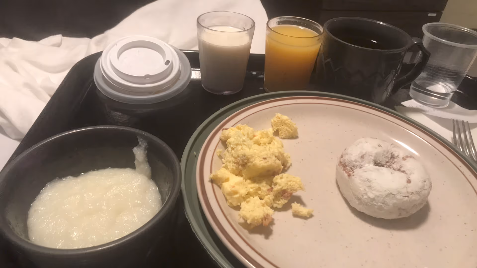 Bedside meal tray with porridge, scrambled eggs, a powdered pastry, juice, coffee, and water.