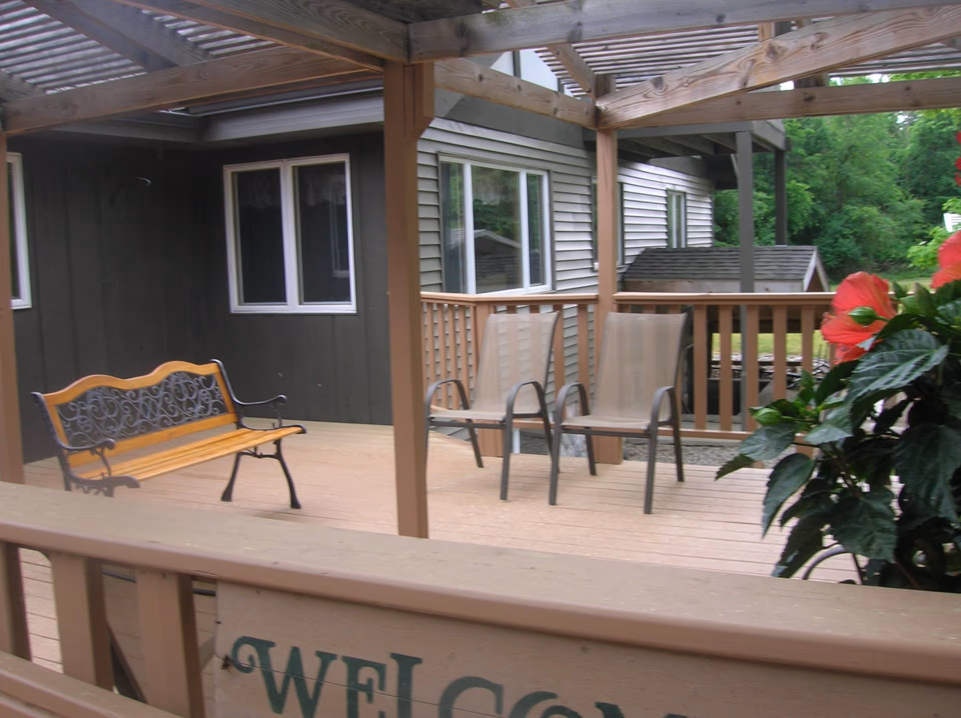 Outdoor wooden deck area with a pergola overhead, featuring a wooden bench with metal armrests and backrest, two beige mesh chairs, and a potted plant with red flowers on the right side. The deck is attached to a building with windows and surrounded by greenery.