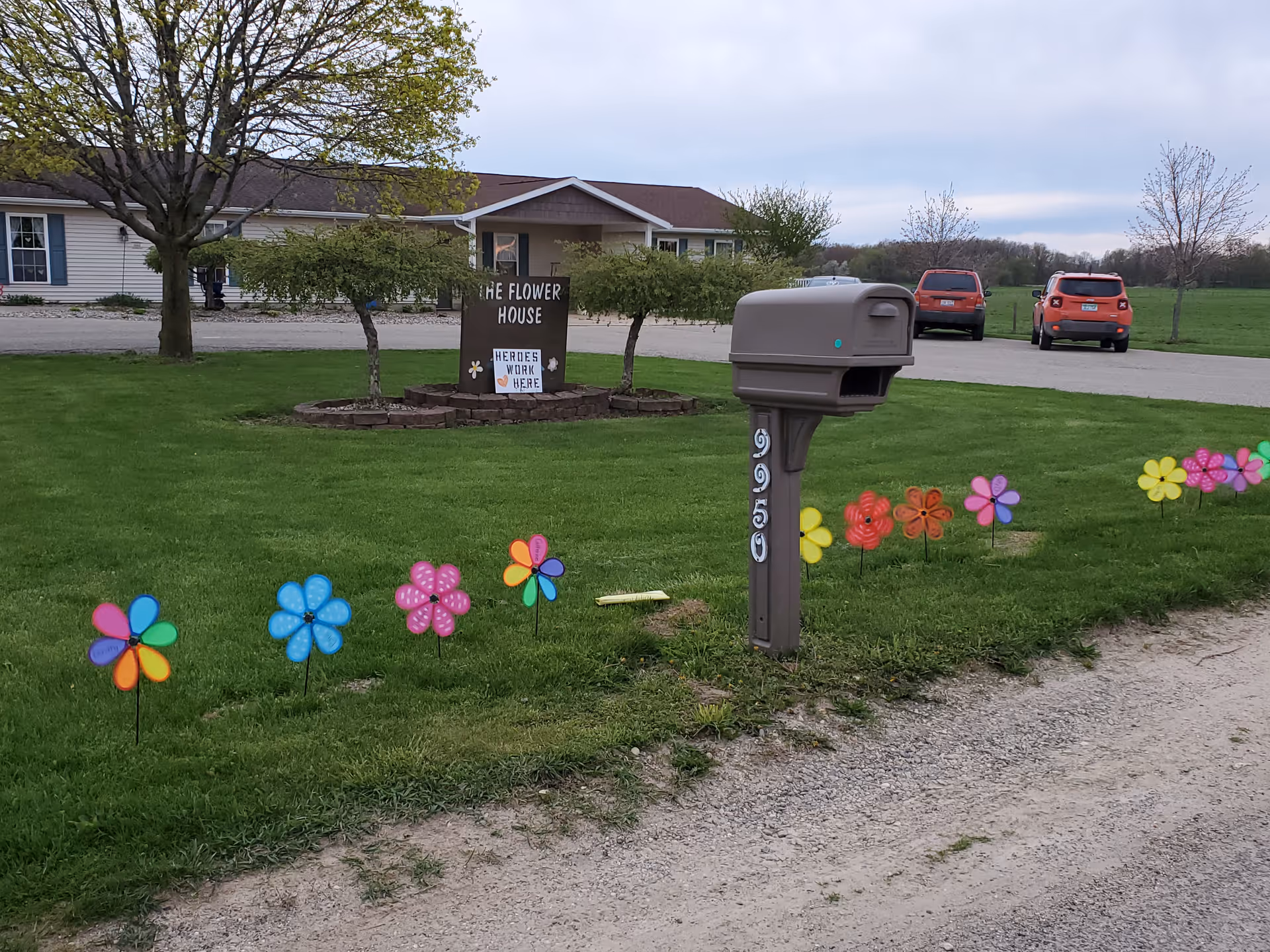 Outdoor view of a facility named The Flower House with a sign that reads 'Heroes Work Here'. The scene includes a mailbox with the number 950, colorful flower-shaped decorations planted in the grass, a gravel driveway, two parked cars, and a building in the background surrounded by trees and green lawn.