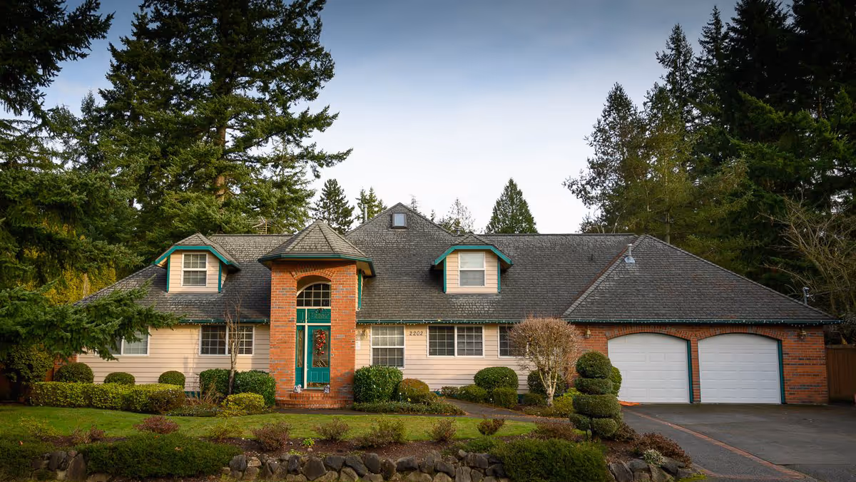Front exterior view of a large, two-story residential building with a brick and beige siding facade, green-trimmed windows, a teal front door with a wreath, and a two-car garage. The house is surrounded by well-maintained landscaping including bushes, trees, and a stone border along the lawn.