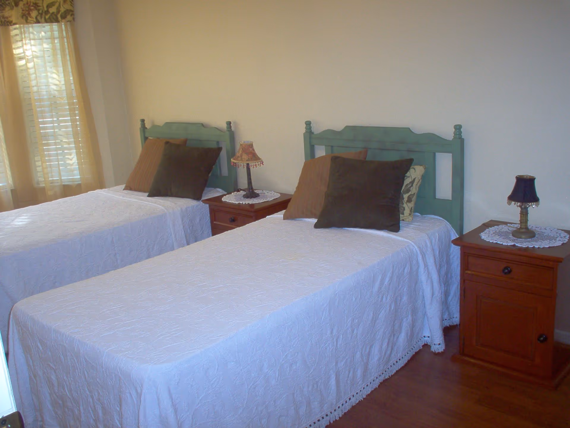 A bedroom with two single beds featuring green wooden headboards, white bedspreads, and multiple pillows. Between the beds are two wooden nightstands, each with a decorative lamp and doily. A window with sheer curtains is visible on the left side of the room.