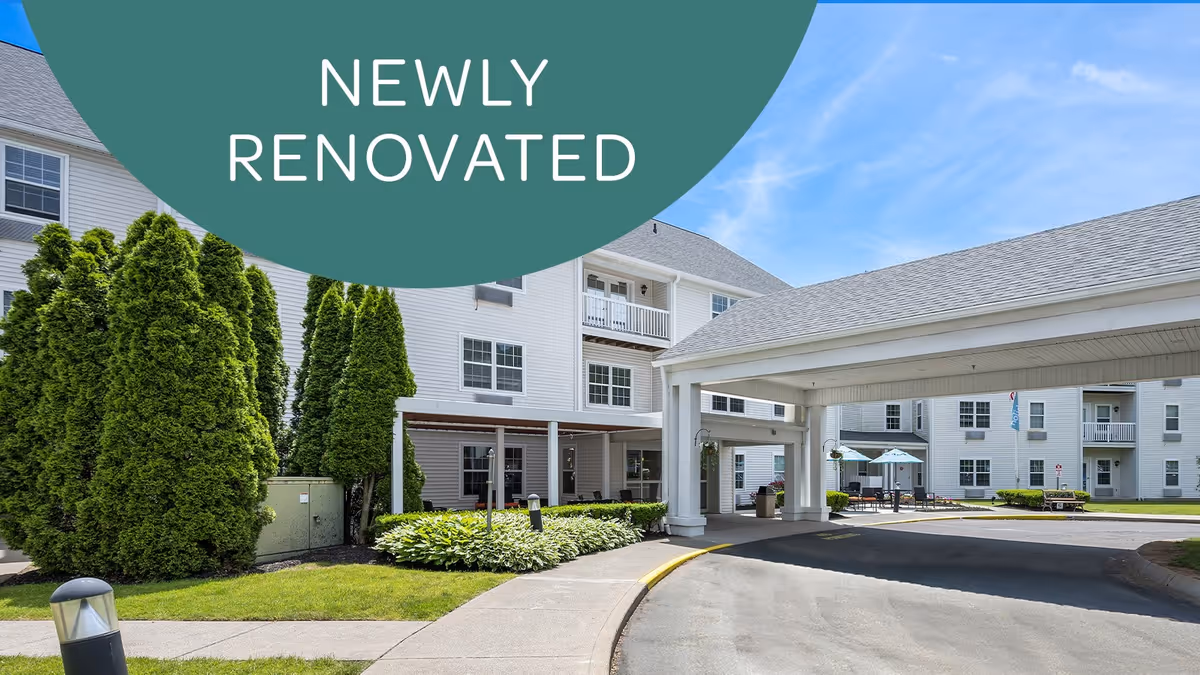 Exterior view of a newly renovated senior living facility with white buildings, a covered driveway entrance, well-maintained landscaping including tall green bushes and a small garden, and outdoor seating with umbrellas under a clear blue sky.