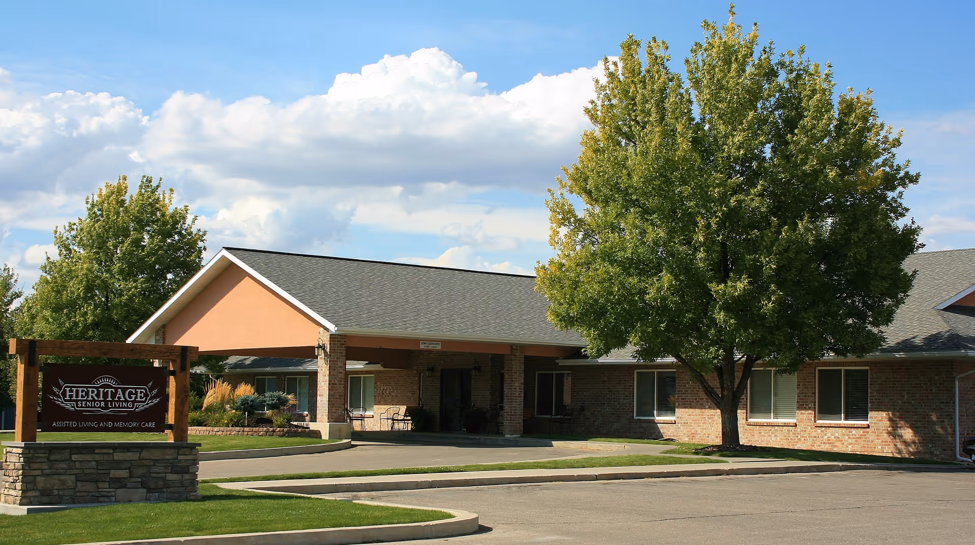 Front exterior of the Heritage Senior Living facility showing the entrance canopy, landscaped sign, and a large tree.