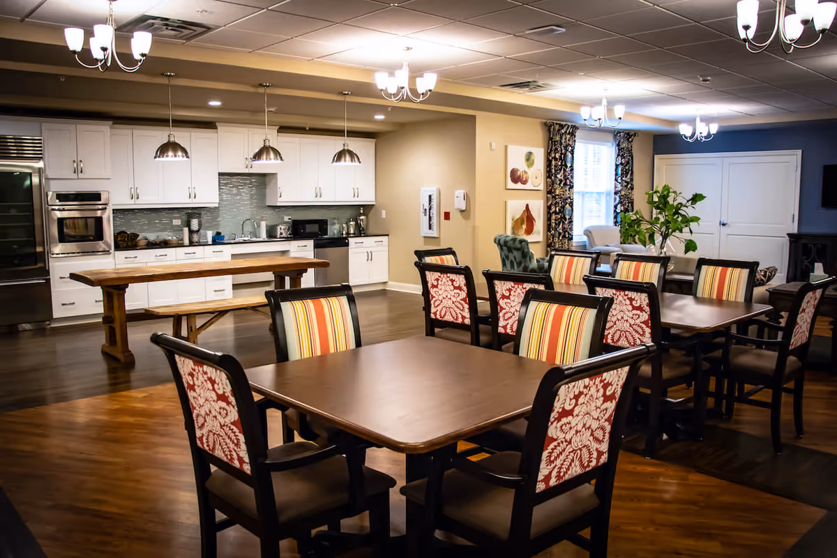 Communal dining area with wooden tables and upholstered chairs facing a modern open kitchen with pendant lighting.