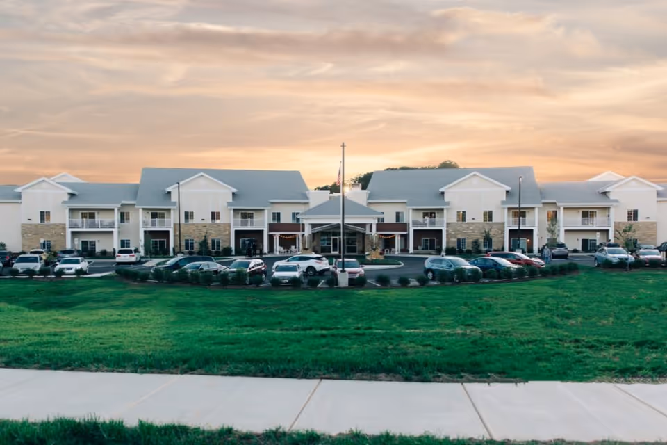 Exterior view of Premier Residences At Tellico Village, a two-story senior living facility with multiple balconies and a covered entrance. Several cars are parked in front of the building, and there is a well-maintained green lawn and sidewalk in the foreground under a partly cloudy sky at sunset.