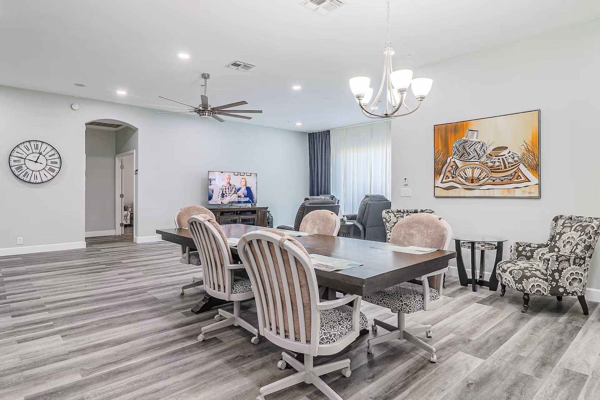 A spacious living room and dining area in an assisted living facility. The room features a large rectangular dining table with six cushioned chairs on wheels. In the background, there are two reclining chairs facing a TV mounted on a wooden stand. The walls are light gray with a large clock on one side and a colorful painting of pottery on another. The floor has gray wood-like planks, and the ceiling has recessed lighting, a ceiling fan, and a chandelier above the dining table. A small side table with a glass top is next to a patterned armchair.