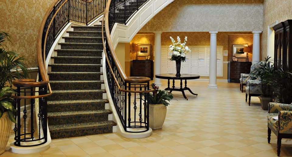 Elegant interior lobby area with a curved staircase featuring a patterned carpet and wrought iron railing. The space includes a round table with a large floral arrangement, potted plants, upholstered chairs, and decorative columns, all set against wallpapered walls with warm lighting.