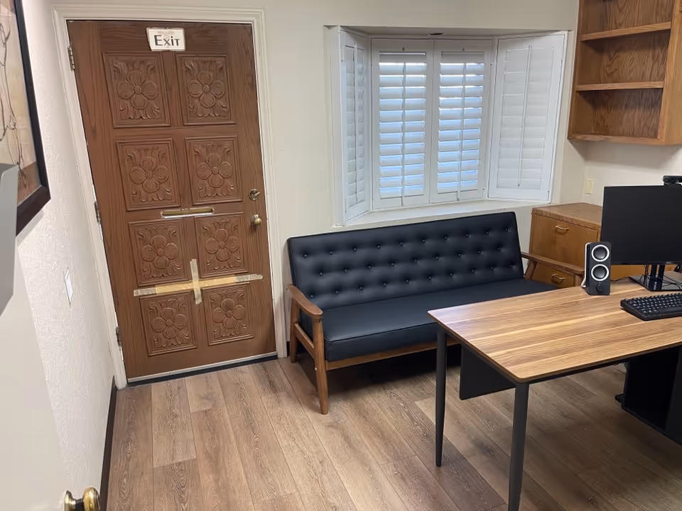 Indoor office area with a black tufted sofa, desk, computer, and window shutters.