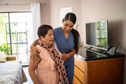 A caregiver in blue scrubs gently supporting and assisting an elderly woman wearing a pink dress and scarf inside a well-lit bedroom with a bed, dresser, and television.
