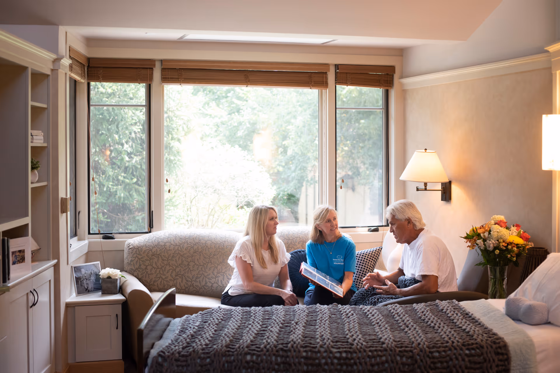 Three people sit on a patterned sofa in a bright hospice living room.