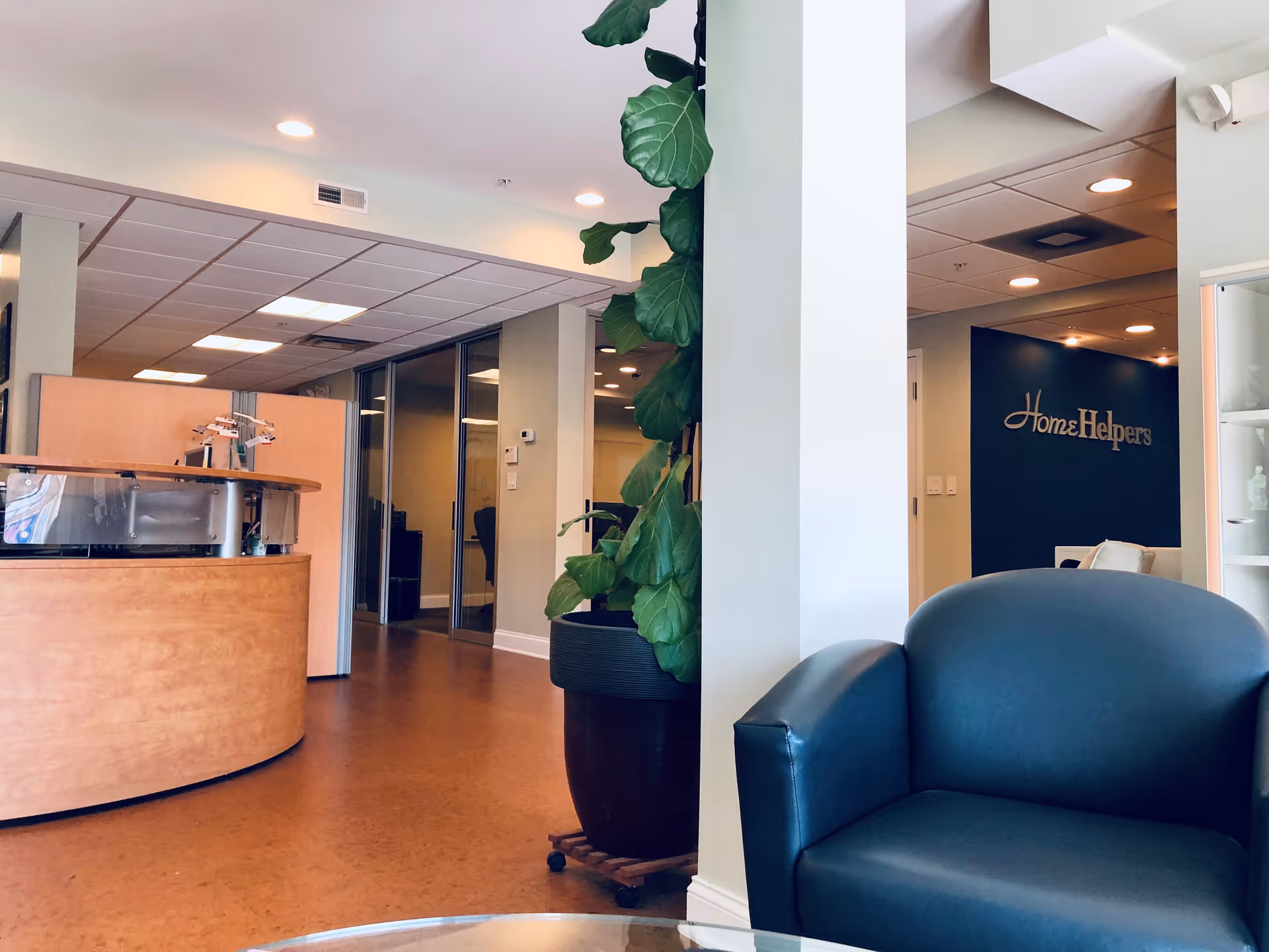 A modern facility lobby with a curved reception desk, plant, and blue Home Helpers sign.