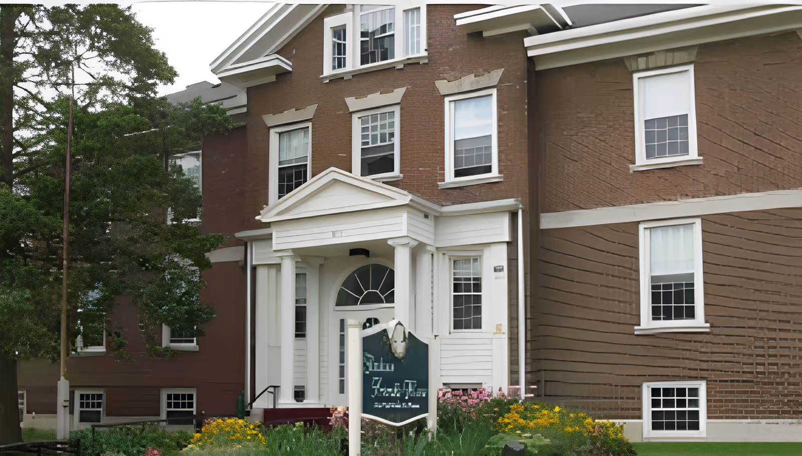Front exterior of a brown brick senior living building with a white columned entrance, landscaping, and a sign near the walkway.