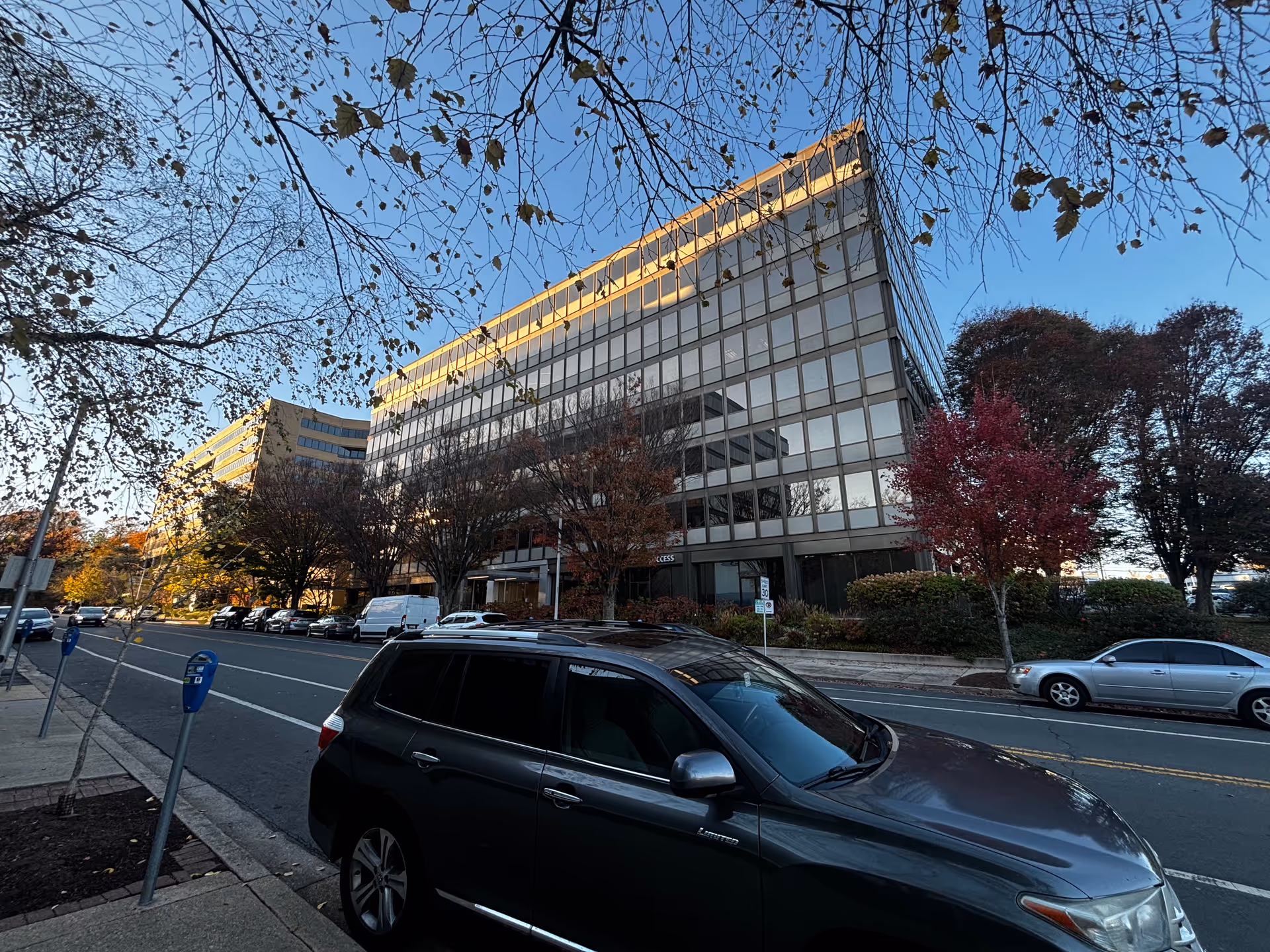 Street view of a multi-story glass-front office building with parked cars and autumn trees.