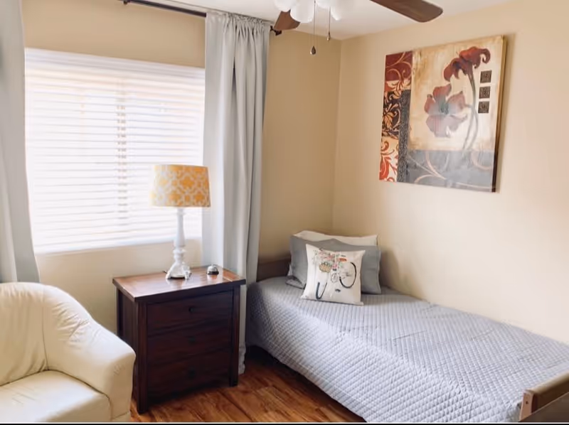 Small furnished bedroom with a twin bed, nightstand and lamp beside a window and armchair.