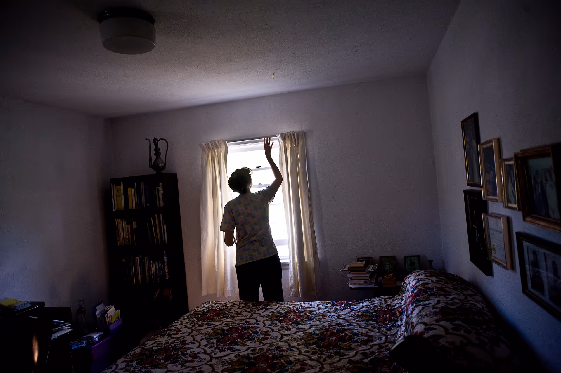 A resident stands by a window in a dim bedroom with a floral bedspread.