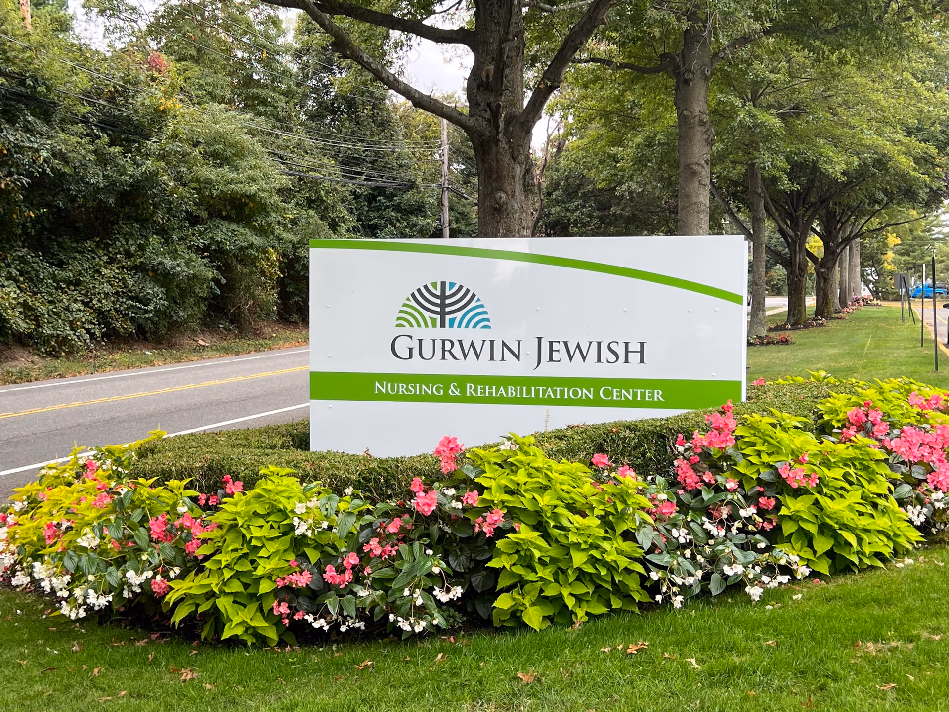 A large white sign with green accents that reads 'Gurwin Jewish Nursing & Rehabilitation Center' surrounded by colorful flowers and greenery, with trees and a road in the background.