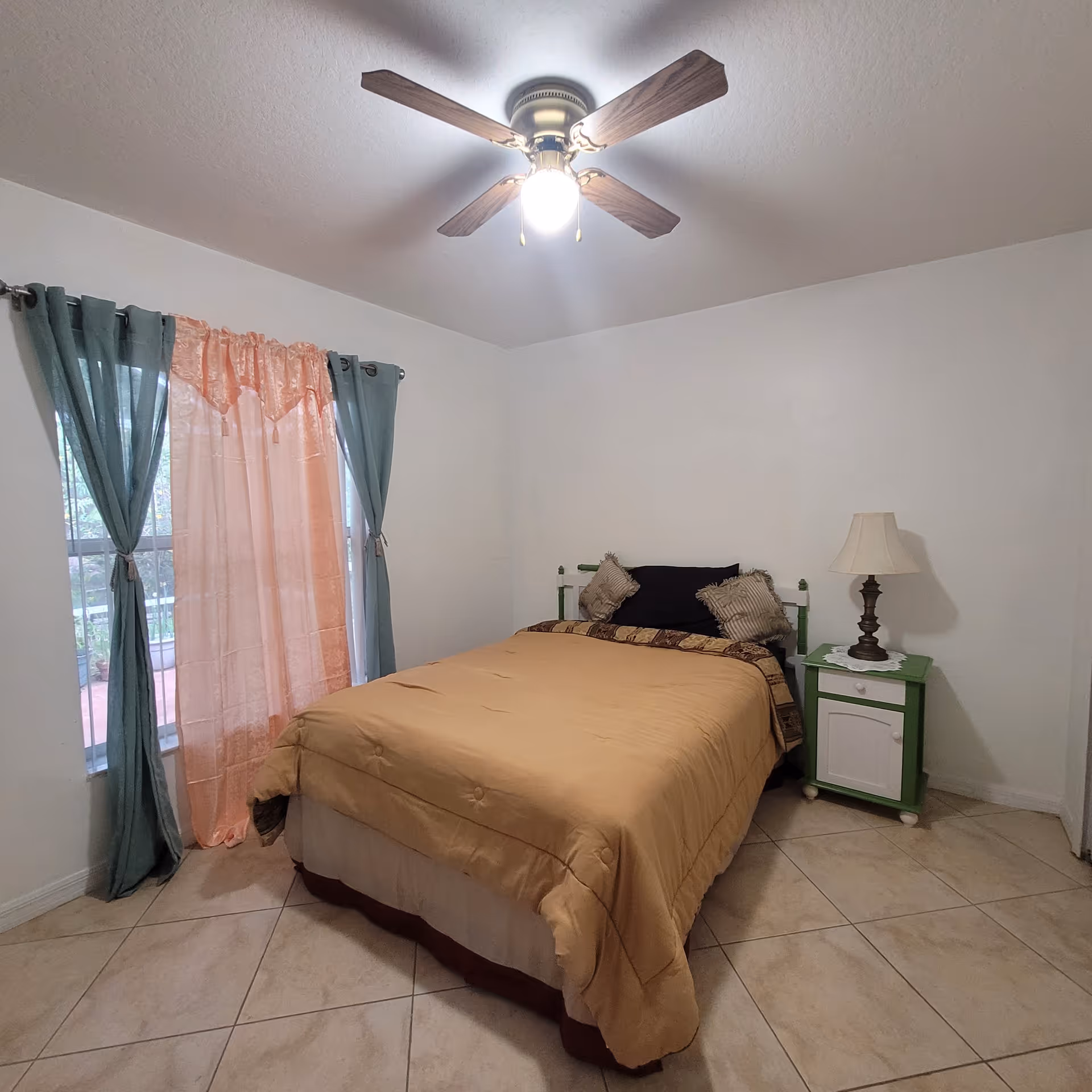 A simple bedroom with a bed covered in a beige comforter and two pillows. There is a green and white nightstand with a lamp on it next to the bed. The room has tiled floors, white walls, a ceiling fan with a light, and a window with peach and teal curtains.