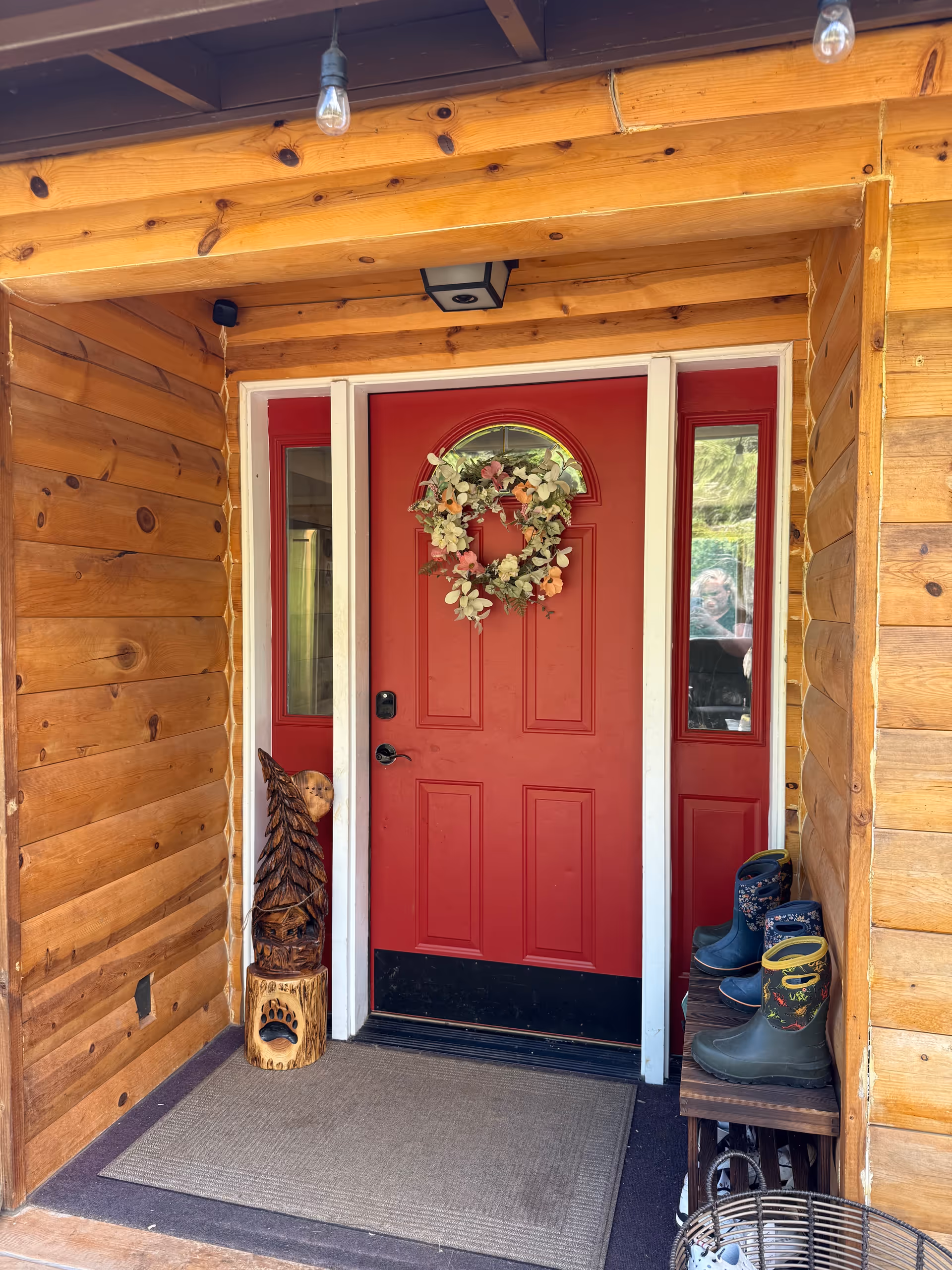 Rustic wood exterior with a red front door, wreath, and boots on the entry path.