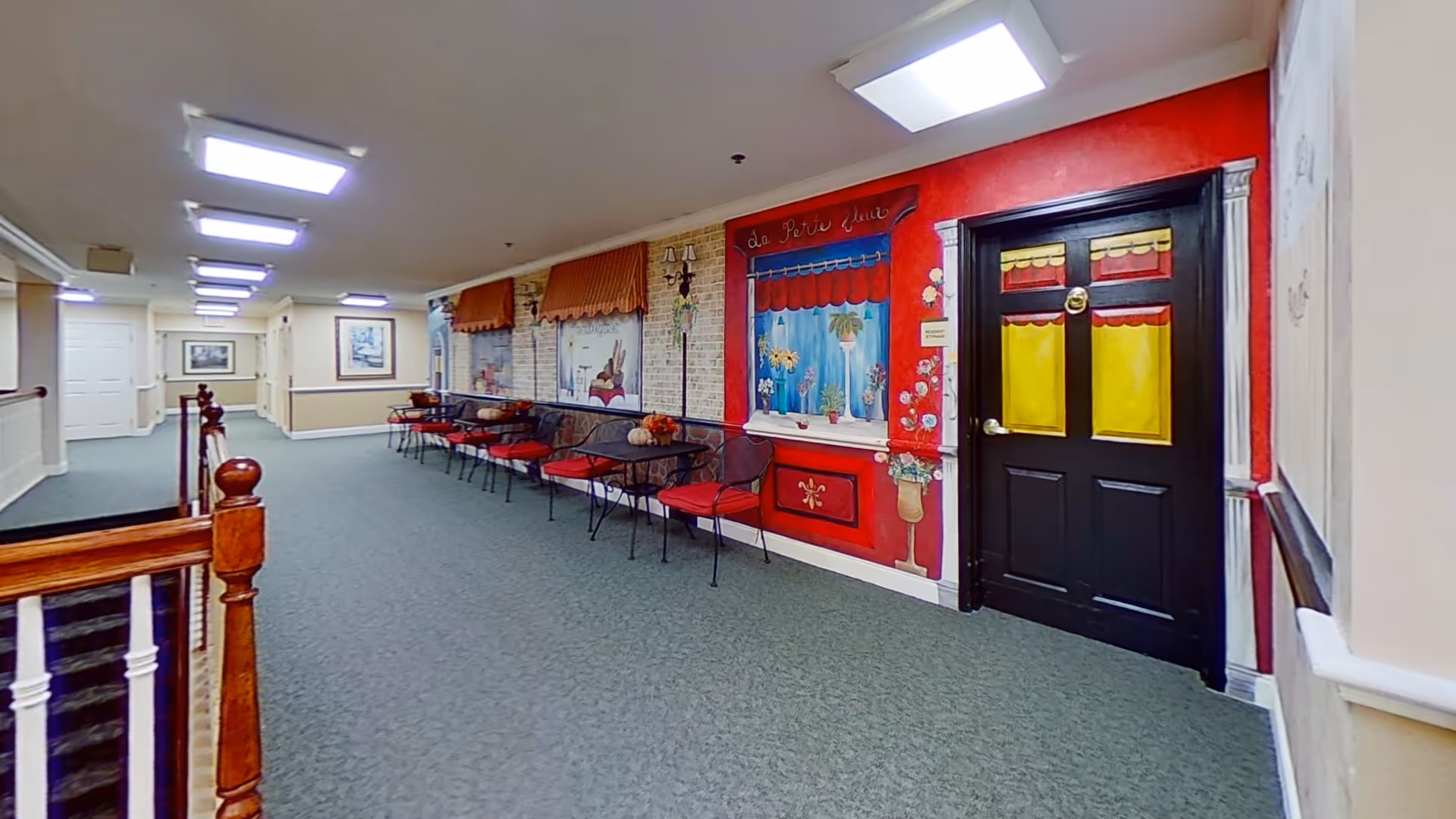 A long indoor hallway with green carpet and white walls, featuring a decorative mural on the right wall that resembles storefront windows and a door painted in bright colors. Several black metal chairs with red cushions and small tables are lined up against the mural wall. The ceiling has multiple rectangular fluorescent lights, and there is a wooden railing on the left side.