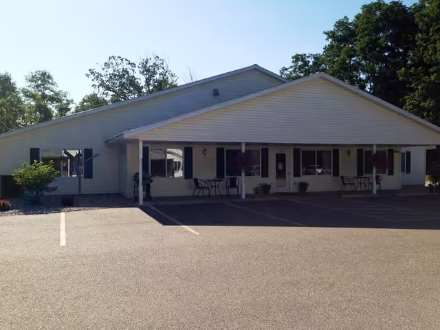 Single-story building with a covered porch, outdoor seating with tables and chairs, and a parking lot in front. The building has light-colored siding and dark shutters on the windows, surrounded by trees and greenery.